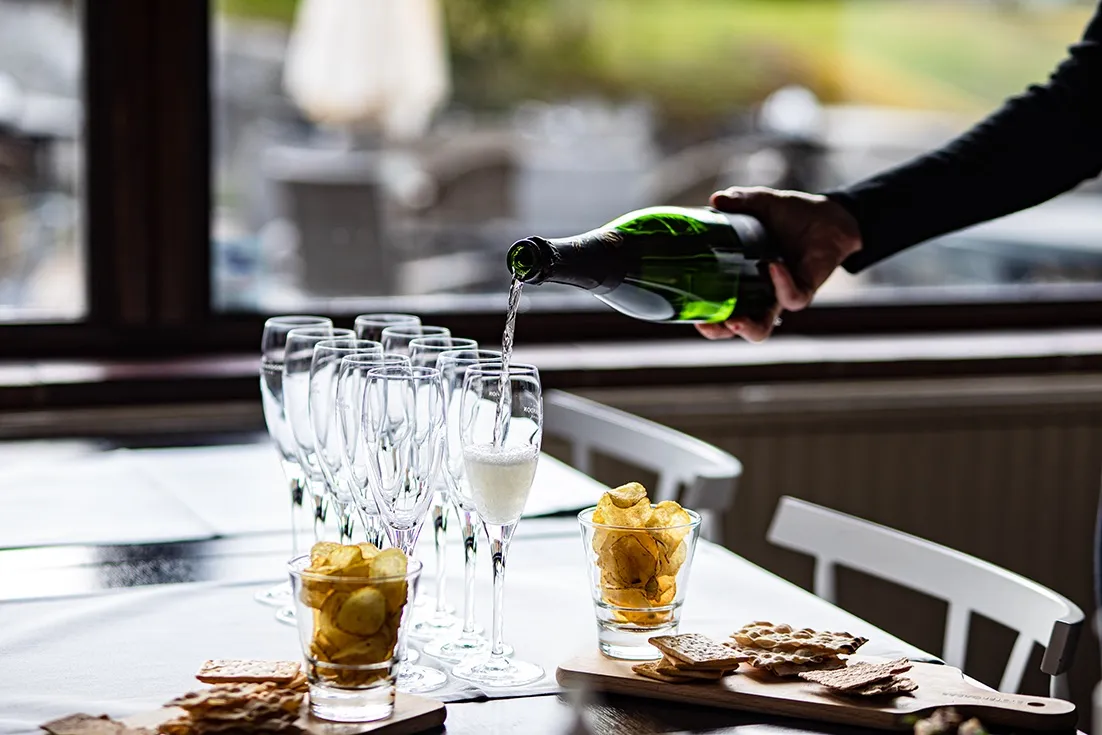 En flaska mousserande vin som häller upp i ett champagneglas, med fler glas i bakgrunden på ett vitt bord.